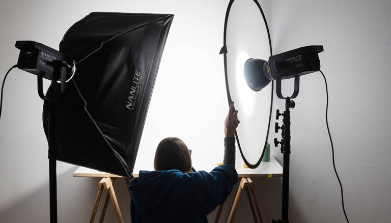 Vista da dietro di una persona all'interno di uno studio fotografico, posizionata davanti a un tavolo da lavoro e circondata da luci professionali: a sinistra c'è un grande softbox Nanlite e a destra un faretto puntato verso un pannello diffusore circolare.