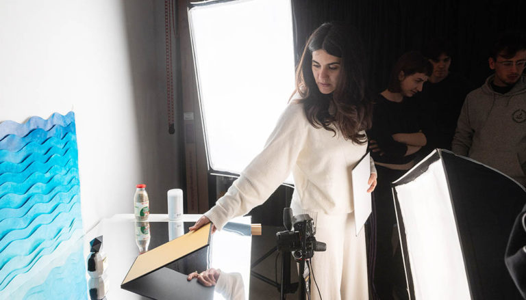 Una ragazza con una maglia bianca allestisce un set fotografico di still life su una superficie specchiante, sistemando un cartoncino dorato vicino a un flacone di profumo; sullo sfondo, altre persone osservano la scena in studio accanto a un grande pannello luminoso.