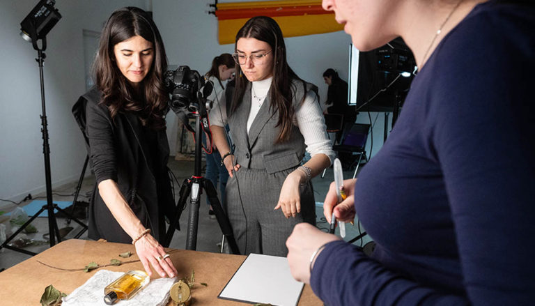 Due ragazze lavorano all'allestimento di uno shooting fotografico su un tavolo in studio, mentre una terza persona in primo piano tiene in mano una matita. Sul tavolo ci sono una bottiglia di profumo, foglie secche e una fotocamera professionale montata su un treppiede Manfrotto.