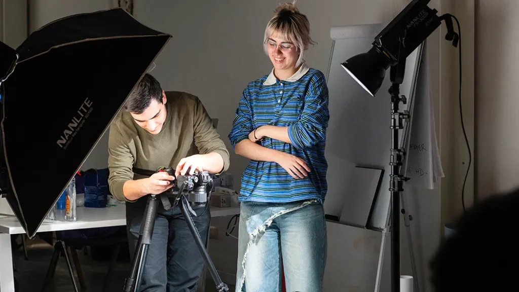 Due studenti durante uno shooting fotografico in studio: un ragazzo regola una fotocamera su treppiede accanto a un softbox Nanlite, mentre una ragazza con maglia a righe osserva sorridendo sotto le luci di scena.