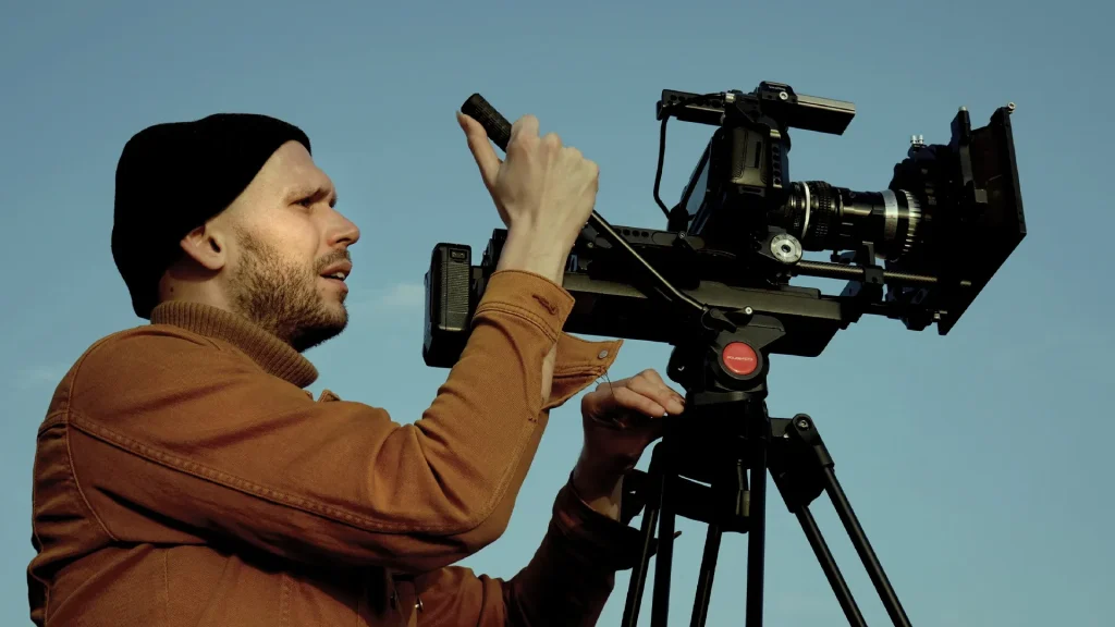 Un operatore cinematografico di profilo con berretto nero e giacca marrone mentre manovra una videocamera professionale montata su un cavalletto, sullo sfondo di un cielo azzurro e limpido.