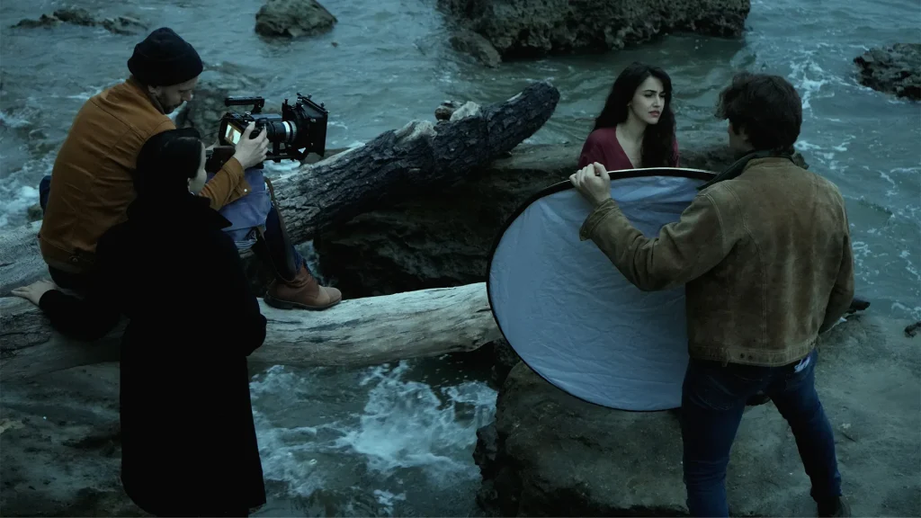 Backstage di un set cinematografico su una scogliera: una troupe composta da tre persone riprende un'attrice con un vestito rosso, mentre un assistente direziona la luce con un pannello riflettente circolare.