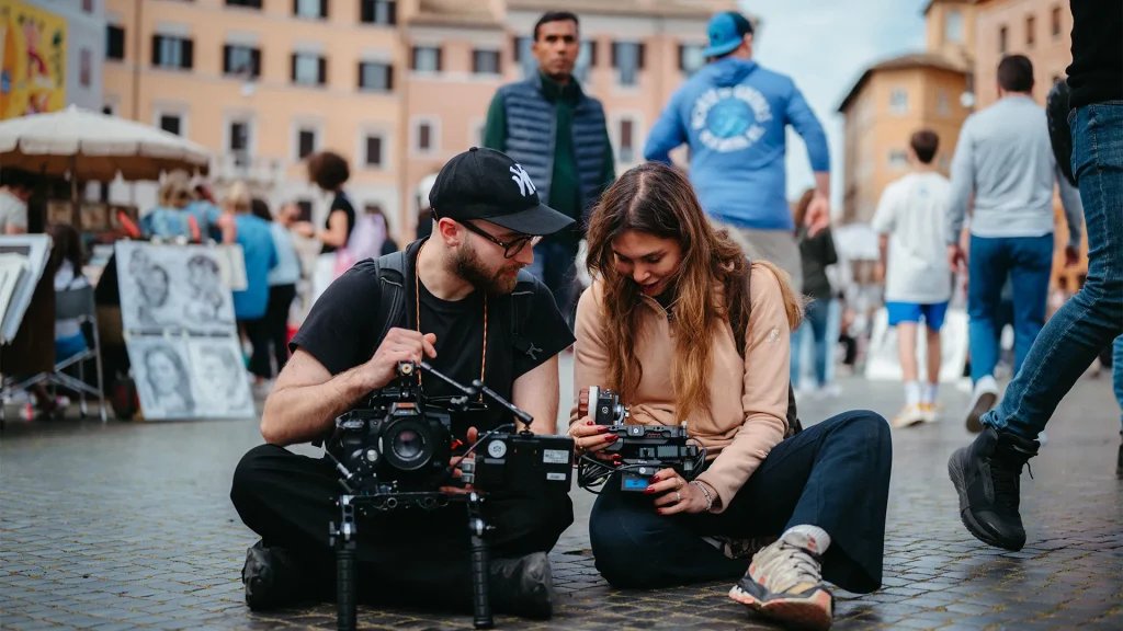 Un uomo con cappellino nero e occhiali e una donna seduti per terra in mezzo a una piazza affollata mentre controllano e regolano due videocamere cinematografiche professionali.