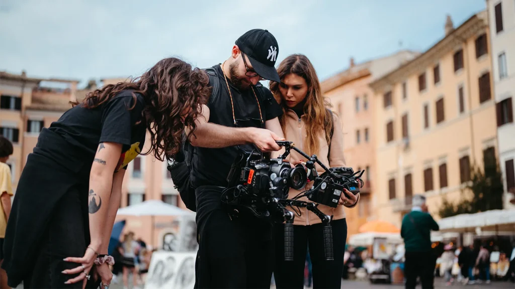 Tre membri di una troupe cinematografica, tra cui un uomo con cappellino NY e una donna con i capelli ricci in primo piano, concentrati nel configurare una videocamera montata su uno stabilizzatore in una piazza cittadina.