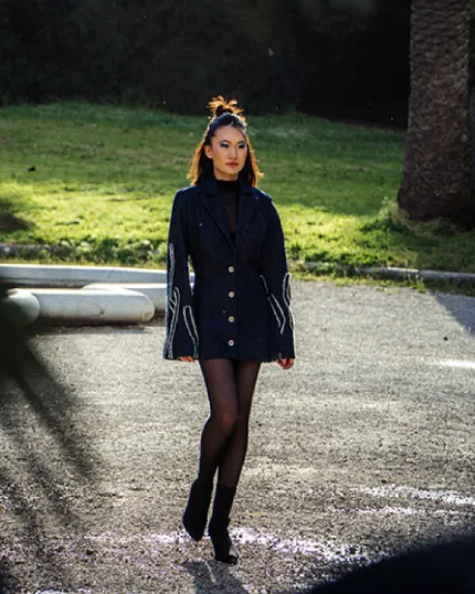Modella con acconciatura raccolta che sfila all'aperto indossando un abito corto scuro stile giacca con maniche ampie e ricamate.