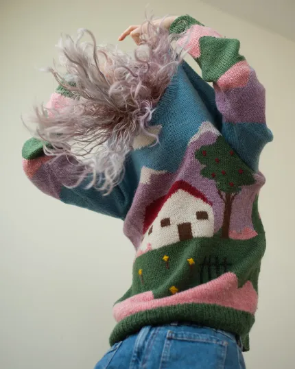 Persona di spalle con capelli mossi sfumati color lilla, mentre indossa un maglione artigianale a maglia con il disegno di una casa in un paesaggio.