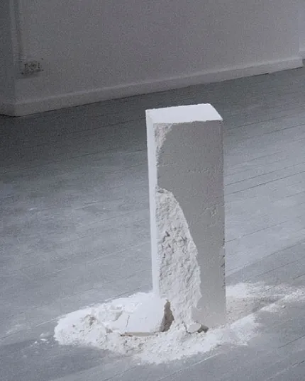 Blocco di gesso bianco a base quadrata parzialmente scolpito o scheggiato, posizionato sul pavimento grigio di un atelier con polvere e frammenti alla base.