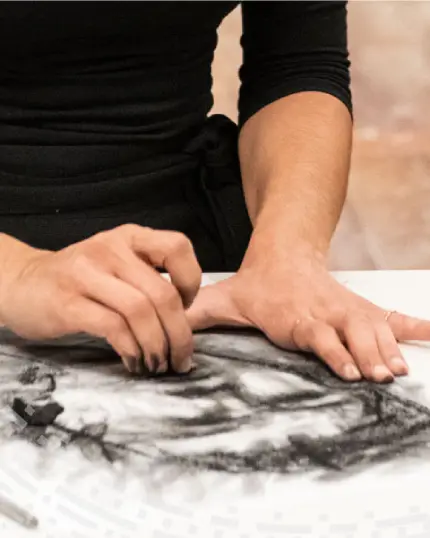 Mani di un artista che lavorano a un disegno anatomico sfumando il carboncino scuro sulla carta bianca.
