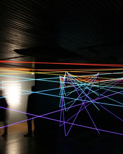 Un'installazione artistica immersiva al buio, caratterizzata da una fitta rete di fili o raggi luminosi al neon che si incrociano nei toni del rosso, giallo, azzurro e viola, con ombre sfocate di visitatori sullo sfondo.