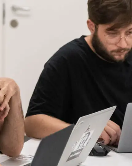 Un ragazzo con gli occhiali e la barba concentrato mentre lavora al proprio computer portatile durante una sessione di formazione in aula.