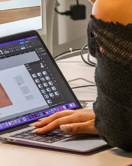 Inquadratura ravvicinata di una mano con unghie curate che utilizza la tastiera di un computer portatile, sullo schermo è visibile l'interfaccia di un software di impaginazione grafica ed editoriale.
