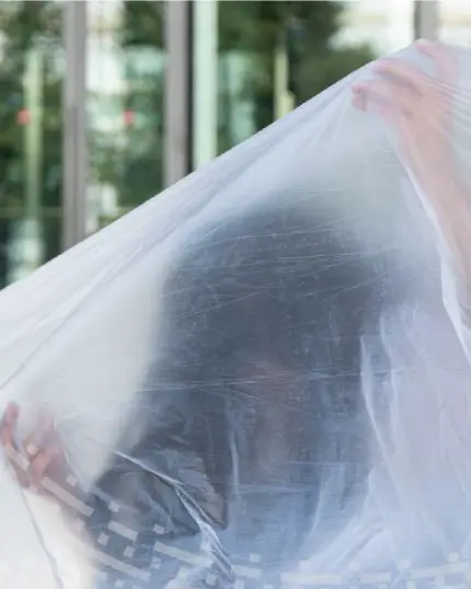 Una persona parzialmente visibile dietro a un grande telo di plastica trasparente e semitrasparente, catturata durante un'azione di performance artistica contemporanea.