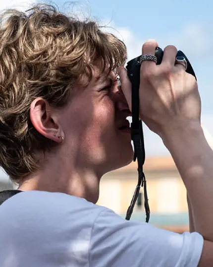 Profilo ravvicinato di un giovane fotografo con i capelli mossi mentre guarda nel mirino della sua fotocamera digitale tenuta saldamente con la mano destra.