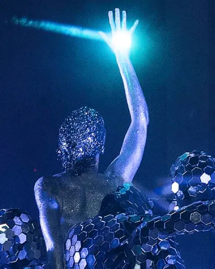 Performance artistica con un performer ricoperto di glitter solleva la mano verso un fascio di luce azzurra - Corso di Scenografia.