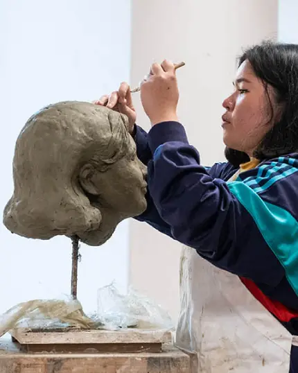 Una studentessa di profilo concentrata mentre modella con una stecca da scultura un busto in argilla che raffigura una testa femminile posizionata su un apposito supporto da officina d'arte.
