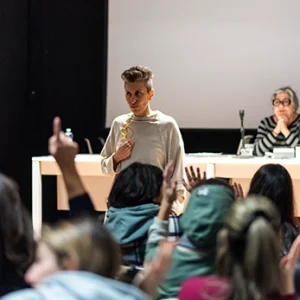 Una relatrice con i capelli corti parla davanti a un pubblico in un'aula universitaria o una sala conferenze, mentre alcuni studenti in primo piano alzano la mano per fare domande.