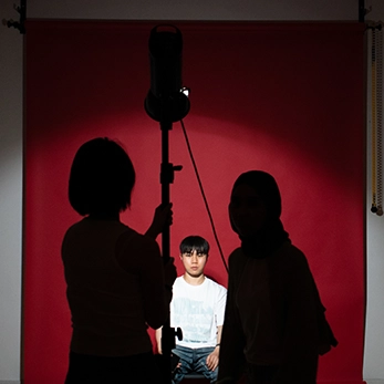 Studio fotografico con sfondo rosso: un ragazzo in maglietta bianca posa seduto, illuminato da un faretto, mentre due figure in primo piano appaiono in silhouette mentre gestiscono le luci e l'attrezzatura.