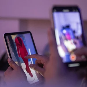 Dettaglio delle mani di una persona che utilizza un tablet per interagire con il modello 3D di un abito rosso, mentre un'altra persona sullo sfondo registra la scena con uno smartphone.