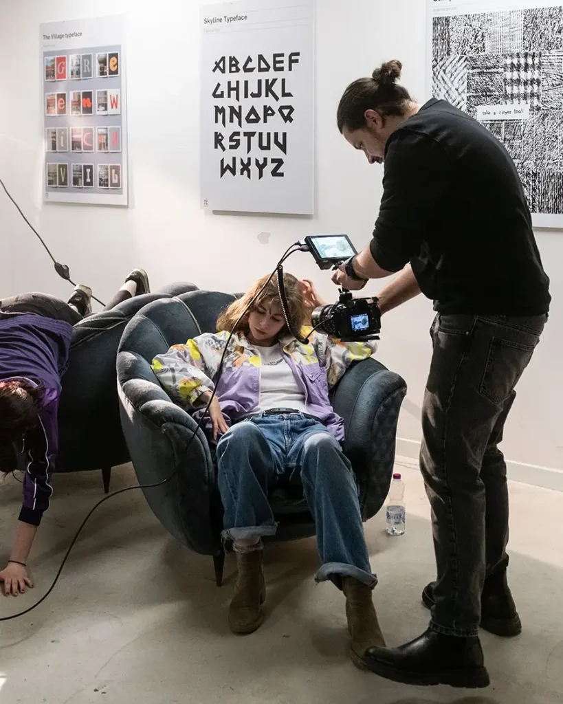Un operatore video riprende con una telecamera a mano una ragazza seduta su una poltrona di velluto blu in un interno con poster di font tipografici alle pareti.