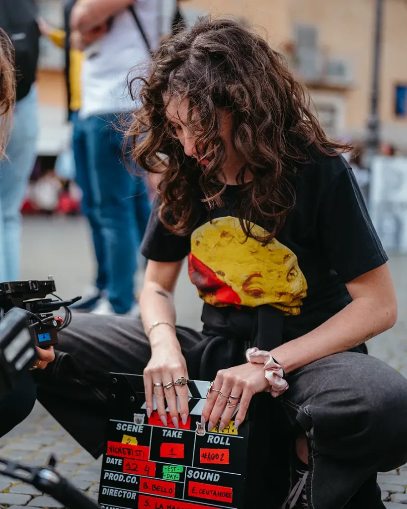 Una ragazza con i capelli ricci e una maglietta nera seduta a terra mentre prepara un ciak cinematografico compilato a mano durante una produzione all'aperto.