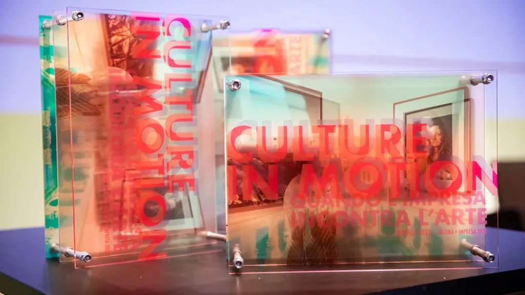 Due targhe trasparenti in plexiglass con bulloni metallici agli angoli, sovrapposte a fotografie artistiche. Sulla targa in primo piano è stampato il testo rosso sfumato "CULTURE IN MOTION - QUANDO L'IMPRESA INCONTRA L'ARTE - MONICA SILVA - CULTURA + IMPRESA 2024".