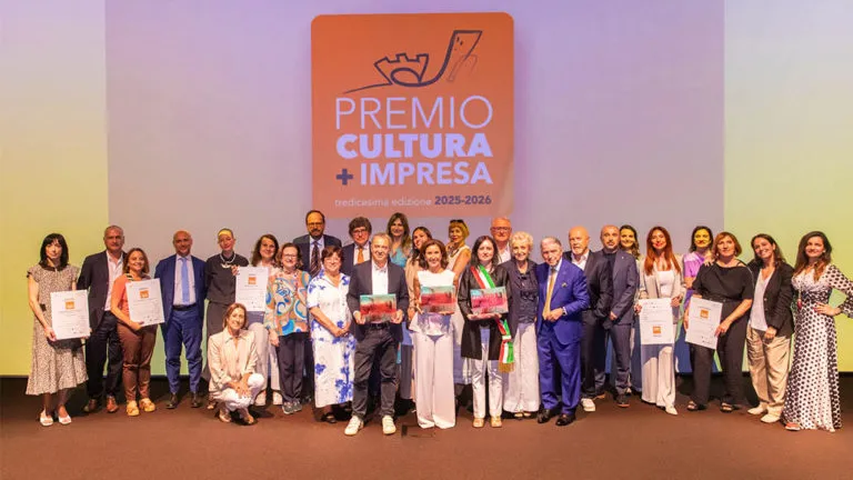 Foto di gruppo dei vincitori e dei partecipanti sul palco per la tredicesima edizione del Premio Cultura + Impresa 2025-2026. Al centro alcune persone mostrano targhe e attestati, mentre sullo sfondo campeggia un grande logo arancione dell'evento.
