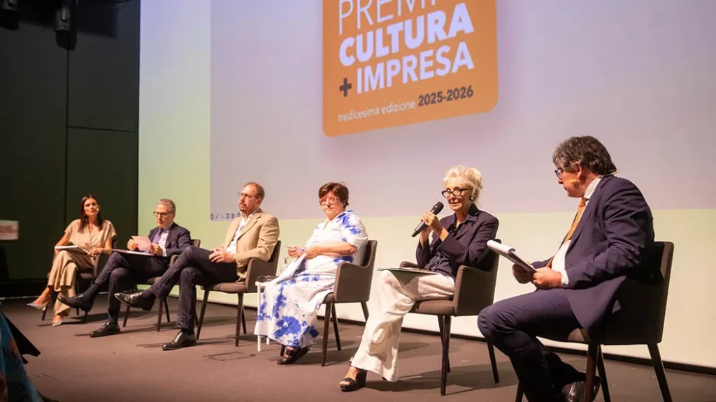 Cinque relatori seduti in poltrona sul palco durante un talk show. Una donna al centro parla al microfono, mentre l'uomo a destra consulta dei fogli. Sullo sfondo è proiettato il logo arancione del Premio Cultura + Impresa 2025-2026.